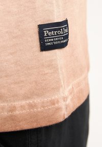 Světlé růžové bavlněné tričko s modrou nálepkou s bílým textem, šitými detaily na lemu a kontrastní černou částí na spodní straně.