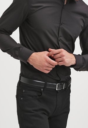 Mężczyzna ubrany w dopasowaną czarną koszulę i czarne jeansy, poprawiający rękawy dłońmi w okolicy talii.