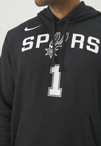 Homme portant un sweat à capuche noir avec un texte blanc "SPURS", le logo de l'équipe au centre, le numéro un dessous et un swoosh blanc Nike sur l'épaule.