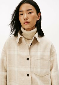 Beige plaid wool shirt jacket with a collar, front pocket, and black buttons, layered over a light beige ribbed turtleneck.