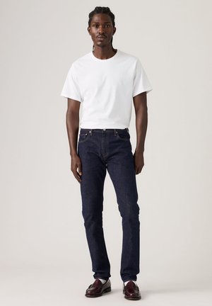 Mężczyzna stoi, ubrany w jednolitą białą koszulkę, ciemnoniebieskie dżinsy i ciemnobrązowe mokasyny na neutralnym tle.