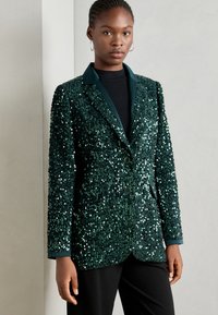 Grønn paljettblazer med reverskrage, to knapper og sidelommer. Strukturert overflate med glatte, reflekterende paljetter.