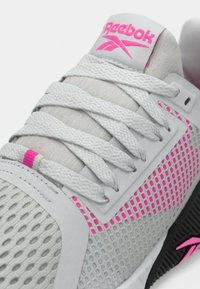 Gaiši pelēki Reebok sniedzēji ar baltām auklām, rozā logo uz mēles, rozā sieta detaļām un melnu zoli ar rozā akcentiem.
