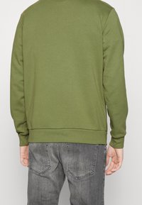 Felpa verde oliva con polsini e orlo a coste, maniche lunghe e texture morbida. Indossata con jeans in denim grigi, vista da dietro.