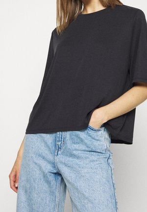 Person trägt ein locker sitzendes schwarzes Kurzarmhemd und hellblaue hoch taillierte Jeans mit einer Hand in der Tasche vor einfachem Hintergrund.