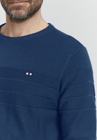 Kék kötött pulóver, amely texturált panelekből, bordás nyakrésszel és egy kis logóval rendelkezik, piros, fehér és kék csíkokkal a mellkason.
