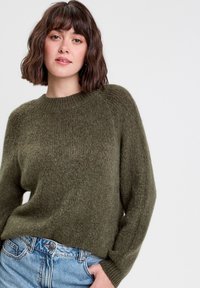 Pull en maille vert olive avec un col rond côtelé et des manches longues, affichant une coupe ample et une texture douce. Associé à un jean bleu clair.