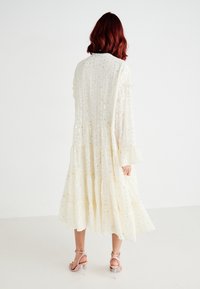 Rochie de culoare crem, cu straturi suprapuse, având o suprafață texturată și paiete împrăștiate. Mâneci lungi cu manșete volan.