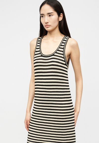 Mlada ženska, oblečena v brezrokavno črno-belo obleko z vodoravnimi črtami, stoji ob enobarvnem svetlem ozadju.