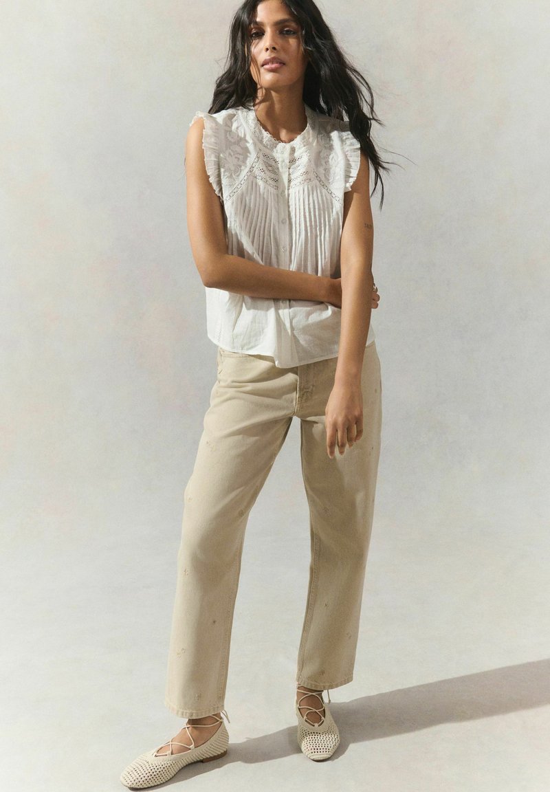 Blouse blanche à manches courtes avec encolure volantée et devant plissé, associée à un pantalon droit beige et des ballerines tissées beiges.