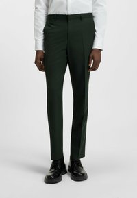 Dunkelgrüne Hosen mit schlanker Passform, hergestellt aus glattem Stoff. Merkmale sind eine flache Vorderseite, Gürtelschlaufen und Taschen. Kombiniert mit schwarzen Dressschuhen.