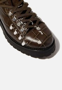 Botín de cuero sintético marrón con textura de cocodrilo, puntera redondeada, superficie texturizada, cordones marrones y herrajes plateados a lo largo de los ojales.