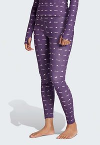 Μωβ κολάν με επαναλαμβανόμενο λευκό λογότυπο και μοτίβο κειμένου. Φτιαγμένα από ελαστικό υλικό, εφαρμόζουν στενά στο σώμα.