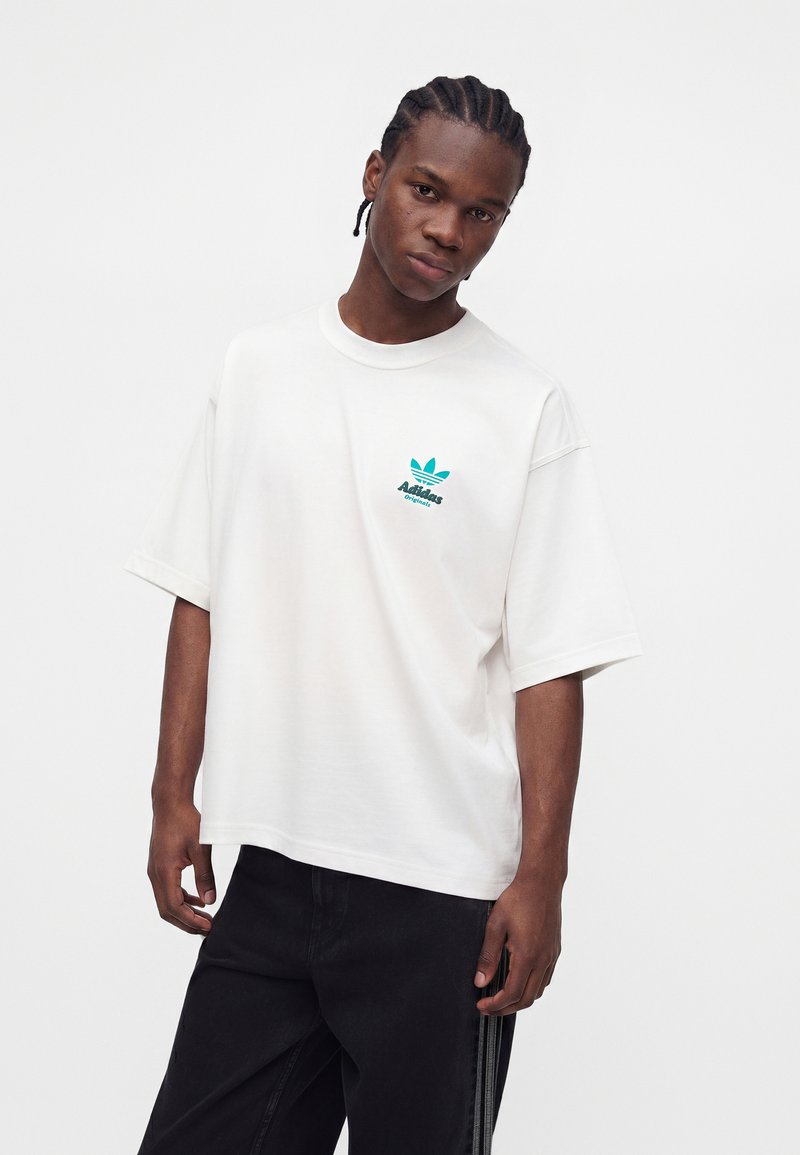 Junger Mann mit geflochtenem Haar, der ein weißes Adidas Originals T-Shirt und schwarze Hosen trägt, steht vor einem schlichten weißen Hintergrund.