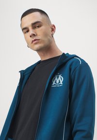 Mėlynas zip-up gobtuvėlis su baltais akcentais ir emblemą kairėje krūtinės pusėje, dėvimas ant juodos marškinėlių su lygia audinio tekstūra.