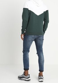 Sudadera con capucha verde y blanca con un diseño de chevrón, combinada con jeans azules desgastados y zapatillas en blanco y negro.