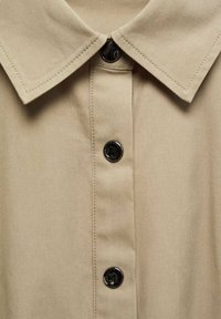 Camicia beige con colletto a punta, dotata di una patta con bottoni neri e una texture di cuciture visibili. Il tessuto appare liscio e robusto.