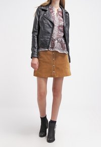 Schwarze Leder-Bikerjacke, gemusterte Bluse, brauner Wildleder-Minirock mit Frontknöpfen, schwarze Ankle-Boots mit Reißverschlüssen, insgesamt geschichteter Look.