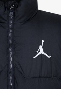 Chaqueta de plumas negra con cuello alto, cierre de cremallera frontal y un logo Jumpman bordado en blanco en el lado izquierdo. Textura suave.