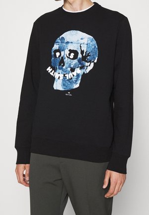 Czarna bluza z niebieskim kwiatowym motywem czaszki i białym napisem "PS Paul Smith" z przodu, noszona z ciemno oliwkowymi spodniami.