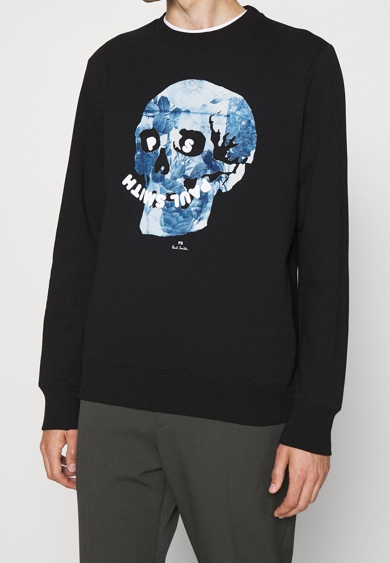 Sweat-shirt noir avec un motif tête de mort florale bleue et texte blanc "PS Paul Smith" sur le devant, porté avec un pantalon vert olive foncé.