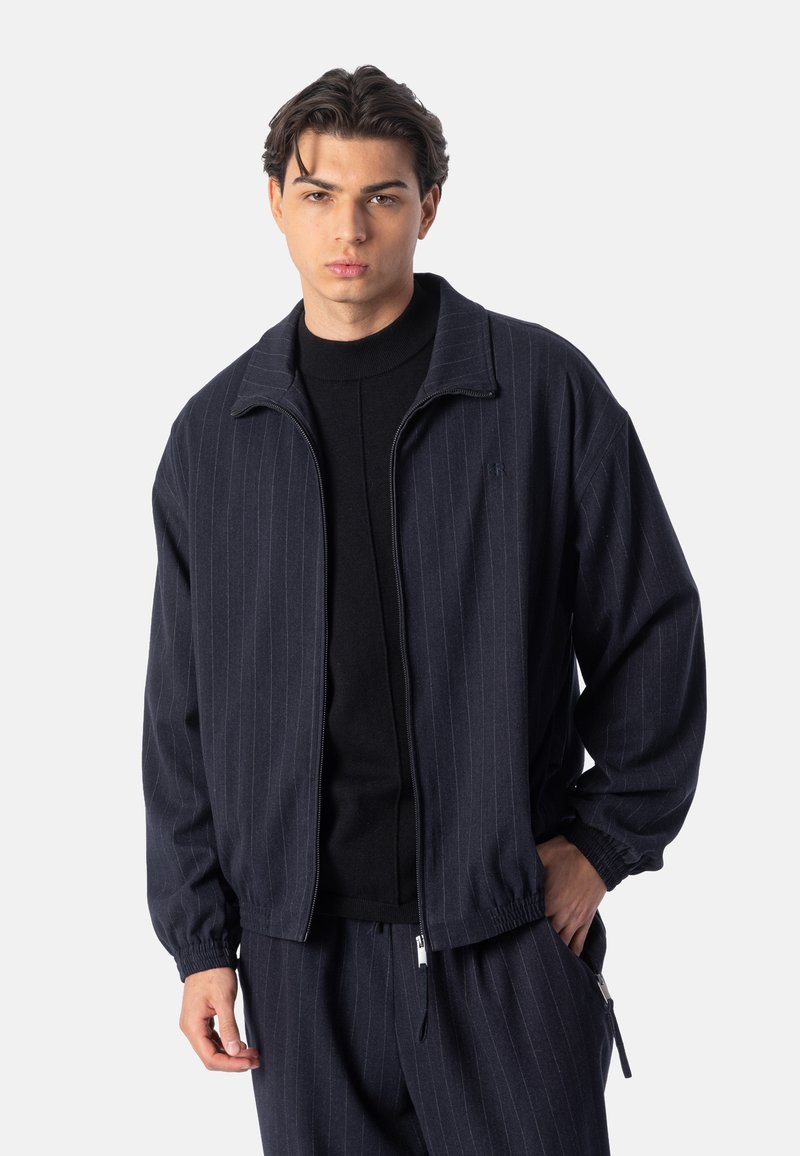Jeune homme portant une veste zippée bleu marine à fines rayures et un pantalon assorti avec une chemise noire en dessous, debout devant un fond uni.
