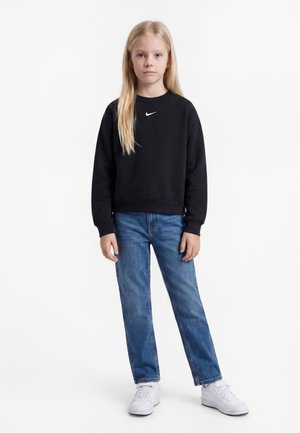 Jeune fille debout, portant un sweat Nike noir, un jean bleu et des baskets blanches, sur un fond blanc uni.