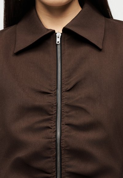 Κοντινή λήψη καφέ ενδύματος με γιακά, με ασημένιο φερμουάρ στο κέντρο και ρυτιδωμένο ύφασμα κατά μήκος της γραμμής του φερμουάρ.