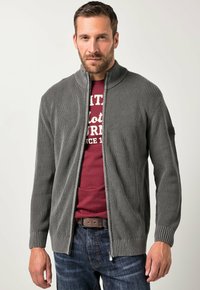 Mann mit kurzen braunen Haaren und Bart, der eine graue Strickjacke mit Reißverschluss über einem roten T-Shirt mit Aufdruck und blauen Jeans vor weißem Hintergrund trägt.