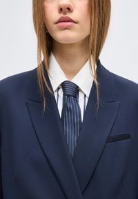 Νεαρός με ανοιχτό καφέ μαλλιά, φοράει σκούρο μπλε σακάκι, λευκό πουκάμισο με μπλε ρίγες και γραβάτα με ταιριαστές ρίγες.