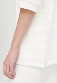 Witte gebreide kleding met een textuurpatroon, voorzien van een split aan de onderkant en een zachte, relaxte pasvorm. Hand aan de zijkant geplaatst.