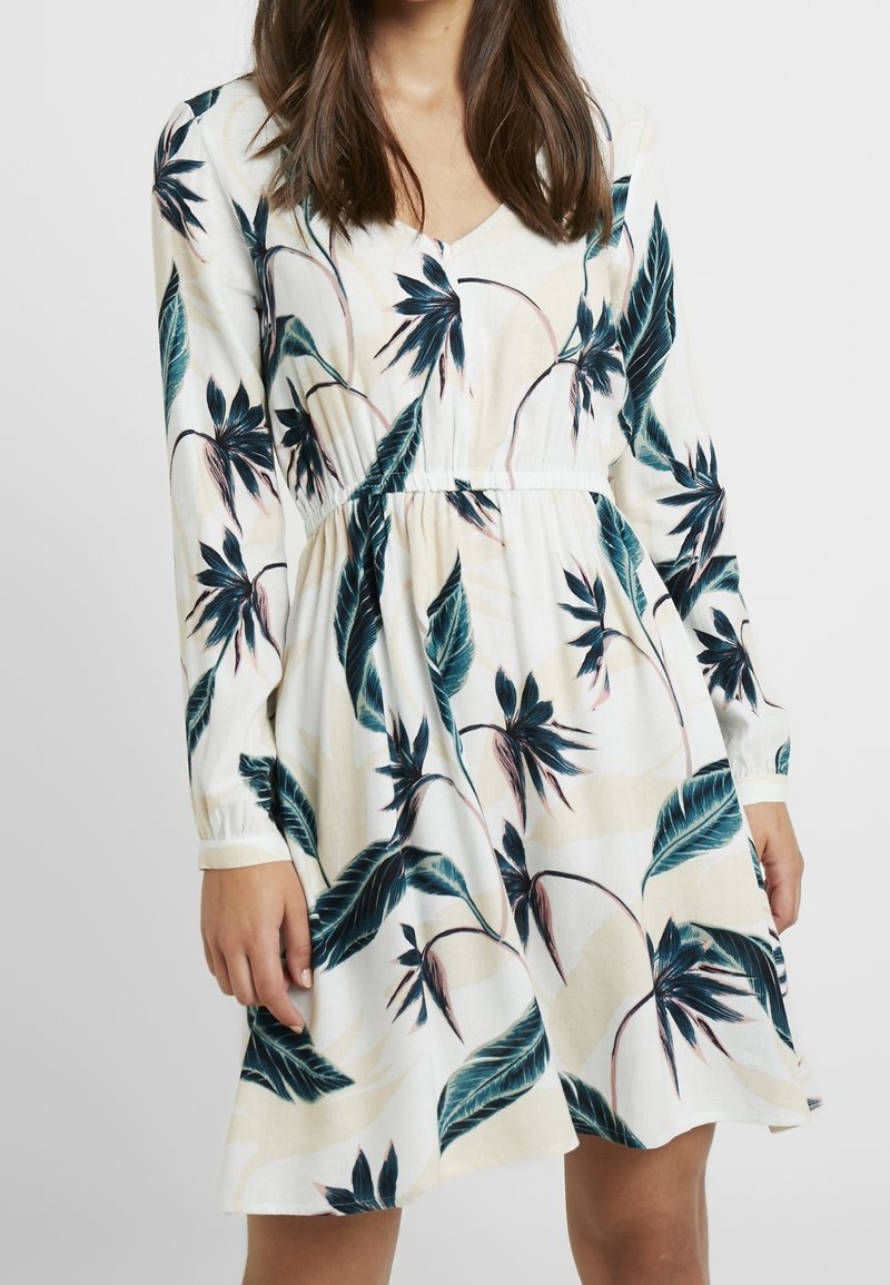 Femme portant une robe blanche à manches longues avec un imprimé de feuilles et fleurs tropicales bleu et vert.