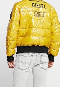 Giacca imbottita gialla con texture lucida, chiusura frontale con zip, polsini neri a costine e dettaglio logo sul retro. Indossata sopra jeans grigio chiaro.