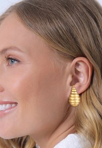 Boucles d'oreilles en ton or en forme de goutte d'eau, avec une texture striée, présentant une finition polie et un attachement de matériel visible.