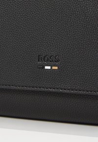 Produit en cuir noir texturé avec logo "BOSS" embosser. Le design comprend trois accents de couleur : noir, blanc et marron.