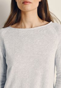 Pruunide pikkade juustega naine, kes kannab heledat halli pika varrukaga särki, millel on lõpetamata äärtega paadi-kaelus, neutraalse taustaga.