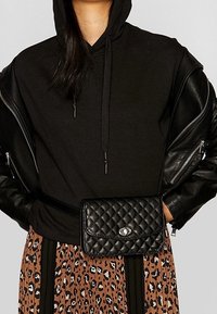 Borsa nera trapuntata con pattina e hardware in argento, indossata su un grande hoodie nero e una gonna leopardata marrone con strisce nere.