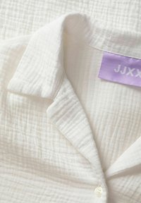 Camisa de algodón blanca con un patrón texturizado y arrugado, con una etiqueta morada y un solo botón blanco en el collar.