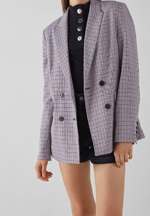 Femme portant un blazer à double boutonnage à carreaux sur un top noir boutonné et un short noir, debout devant un fond clair uni.