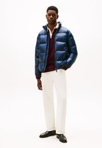 Marineblå pufferjakke med en glansfuld finish, med høj krave og lynlås foran. Parret med en maroon sweater, hvide jeans og sorte sko.