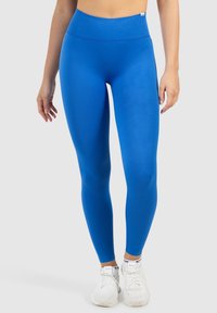 Leggings bleus taille haute avec une texture lisse, dotés d'une large ceinture élastique et d'une coupe ajustée. Associés à des chaussures de sport blanches.