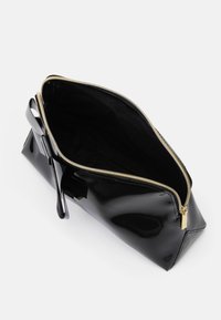 Bolso de charol negro con una cremallera dorada, forma estructurada y un lazo en un lateral. Interior forrado en tela negra, proporcionando un contraste.
