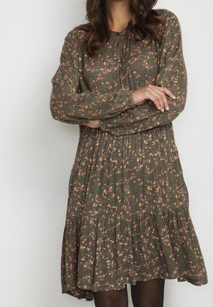 Femme portant une robe à manches longues vert olive avec de petits motifs floraux roses, orange et jaunes, se tenant les bras croisés.