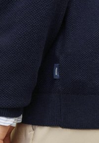 Marinblå stickad tröja med texturerad tyg, med ribbade mudd och fåll, prydd med en liten etikett på sidan. Ljust färgad skjorta synlig under.