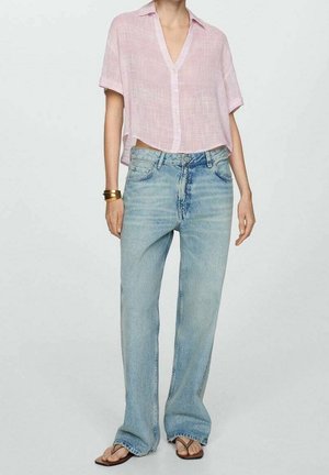 Vrouw draagt een lichtroze kortermige blouse met knopen, lichtblauwe rechte jeans, bruine sandalen en gouden armbanden aan de linkerpols.