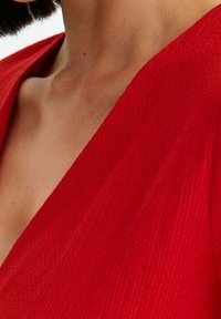 Robe rouge enveloppante en tissu texturé avec un décolleté en V profond. Le tissu présente un motif côtelé subtil, mettant en valeur un design fluide.