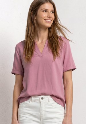 Jeune femme aux longs cheveux bruns souriante, portant un chemisier rose à manches courtes avec un col en V et un pantalon blanc, contre un fond uni clair.