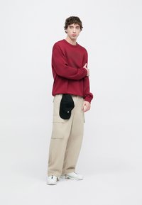 Κόκκινο φούτερ, μπεζ παντελόνι cargo και λευκά αθλητικά παπούτσια. Μαύρο καπέλο κρεμασμένο από το παντελόνι. Το μοντέλο στέκεται μπροστά σε ένα ανοιχτόχρωμο φόντο.