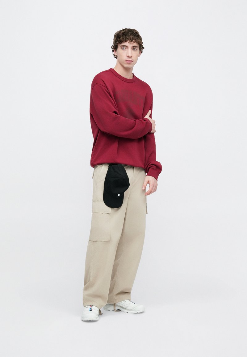 Sweat-shirt rouge, pantalons cargo beige et baskets blanches. Casquette noire suspendue au pantalon. Modèle debout sur un fond clair.