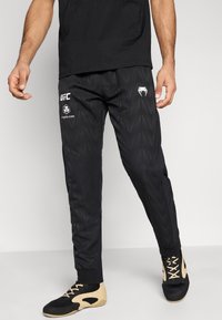 Μαύρο αθλητικό παντελόνι με υφή γεωμετρικού μοτίβου, με λευκά λογότυπα UFC και σχεδιασμένο για εφαρμογή που στενεύει. Φοριέται με μαύρα και μπεζ παπούτσια.